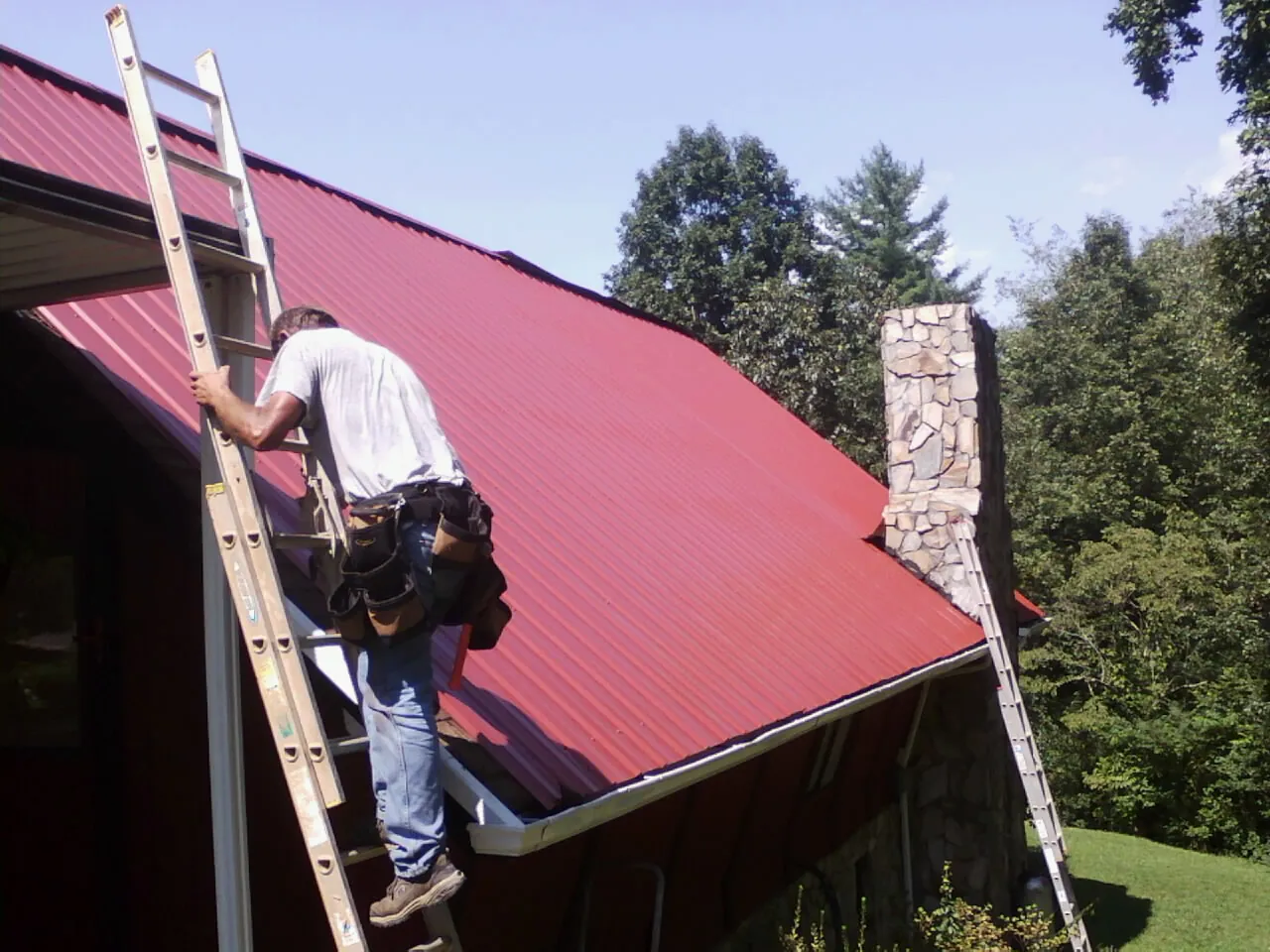 Roofer on a ladder beside a red metal roof and stone chimney — Soffit & Fascia Repair by Roofing Brothers in Millington